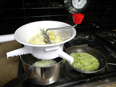 mashing potatoes into the pot with food mill, with green onion mixture in saute pan on the right side.